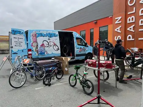 A van with doors open and bicycles around tables outside San Pablo Library