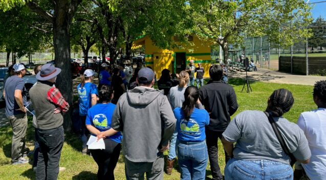 A group of people gathered under trees listening to a speaker 